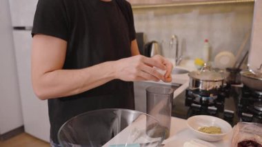 Siyah Tişörtlü Kadın Bir Yumurtayı Bardağa Kırıyor, Mutfak Tezgahı İçi İçi İçi İçi İçi Malzemelerle Dolu, Tencereler Sobada, Beyaz Kaseler, ve Arkaplanda Yemek Utensilleri