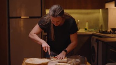 Bir kadın, geceleri bir ev mutfağında, ahşap bir masa üzerindeki pasta katmanını dikkatlice dilimliyor.