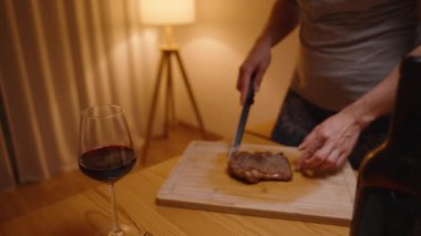 Ahşap bir tahtadan biftek kesen bir kadın. Yanında da bir bardak kırmızı şarap ve sıcak bir yemek.