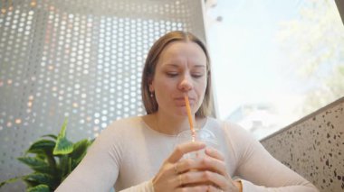 Beyaz kazaklı bir kadın elinde kamışla buzlu kahve tutarken Pepperoni pizzasından bir ısırık alıyor Modern Tasarımlı bir Restoran Masasında oturuyor.