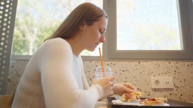 Beyaz kazaklı bir kadın elinde pipetle soğuk bir içecek tutarken pizza ısırıyor arka planda büyük bir penceresi olan bir lokanta masasında oturuyor.