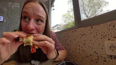 Alüminyum folyo ambalajlı ve buruşuk peçeteli bir restoranda otururken İki Elli Sandviç Tutan ve Büyük Isıran Kadın