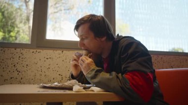 Alüminyum folyo ambalajı ve peçetesiyle Restoran Masasında otururken Kırmızı ve Siyah Ceketli Adam Büyük Bir Sandviç Isırıyor