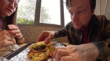 Bir adam restoranda oturmuş, bir kadın onu izlerken sandviçini yiyor, önünde alüminyum folyoyla tepsiyi sarıyordu.