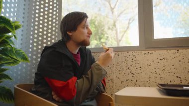 Kırmızı ve Siyah Ceketli Adam Restoran Masasında oturmuş Düşünceli bir ifadeyle Pencereden dışarı bakarken Sandviç yiyor