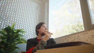 Kırmızı ve siyah ceketli bir adam, bir lokanta masasında pencereden dışarı dikkatlice bakarken elinde bir parça ekmek tutuyor.