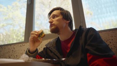 Kırmızı ve siyah ceketli bir adam sandviç tutuyor ve pencerenin yanındaki bir restoranda otururken bir ısırık alıyor.