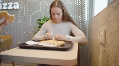 Beyaz kazaklı bir kadın restoran masasında oturmuş önünde siyah bir tepsiyle peynirli ekmek yiyor.