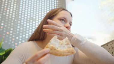 Beyaz kazaklı bir kadın elinde bir dilim peynirli pizza tutuyor ve tatmin olmuş bir ifadeyle ona bakıyor.