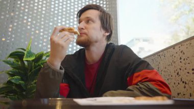 Kırmızı ve Siyah Ceketli Adam Bir Restoranda Pencere kenarında Otururken Pizza Dilimi Yiyor