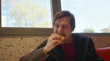 Siyah ceketli ve kırmızı gömlekli bir adam Fast Food restoranında sandviçi ısırıyor. Odaklanmış bir ifadeyle, dışarıda doğal ışık ve bulanık ağaçlarla bir pencerenin yanında oturuyor.