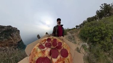 Okyanusun önünde elinde iki dilim Pepperoni Pizza tutan bir kadının yakın çekimi. Güneş gözlüğü ve ekose ceket takıyor.
