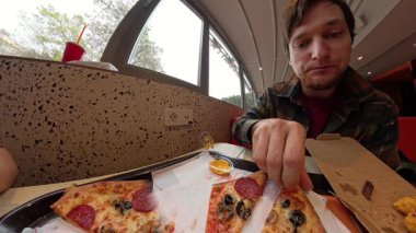 Restoran masasında bir tepsi pizza ve bir sos kabıyla otururken elinde bir dilim Pepperoni Pizza tutan adam geniş açılı bir perspektiften çekilmiştir.