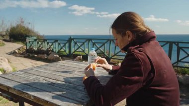 Açık Gök Altında, Okyanus 'un Yakınındaki Ahşap Piknik Masasında Mandalina soyan Siyah Ceketli Bir Kadın Görünümü