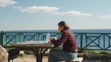 Piknik masasında tek başına oturan bir kadın Mandalina soyuyor Okyanus manzaralı ve açık mavi gökyüzü