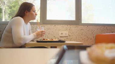 Beyaz kazaklı bir kadın restoranda bir tepsi pizzayla otururken pipetle soğuk bir içecek içiyor. Önü bulanık ve arka planda büyük bir pencere var.