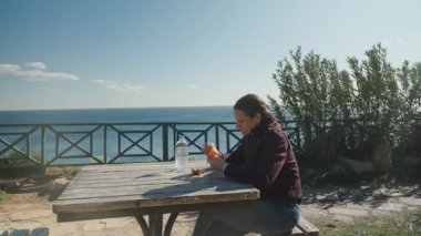 Ahşap piknik masasında oturan bir kadın arkasında bir şişe su ve arka planda okyanusla mandalina soyuyor.