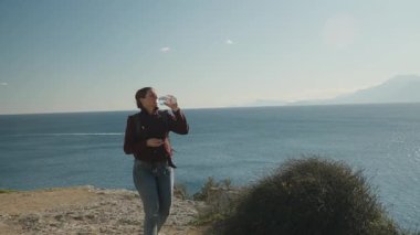 Uçurumun kenarında duran bir kadın Uzaktaki Dağlarla okyanusu seyrederken bir yandan da şişeden su içiyor.