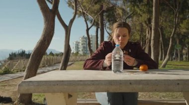 Beton bir piknik masasında oturan, yanında bir şişe su ve mandalinayla çikolatalı çörek yiyen bir kadın.