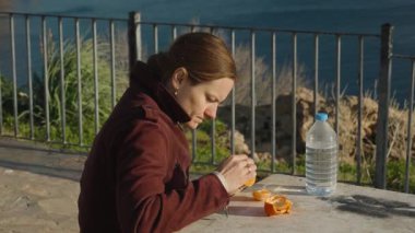 Gün ışığı yüzünü aydınlatırken bir masa başında oturmuş bir şişe suyla mandalina yiyor.