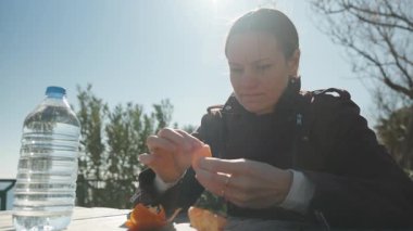 Ahşap bir masada mandalina soyan bir kadın. Güneş ışığı ellerinde parlıyor ve yakınlarda bir şişe su var.