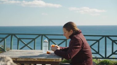 Ahşap bir masada oturan, su şişesiyle mandalina yiyen ve arka planda okyanus manzaralı bir kadın.