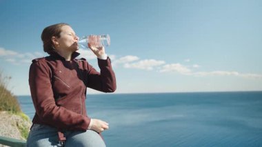 Güneşli bir günde, Çitin üzerinde oturmuş, okyanus manzaralı şişeden su içen kadın.