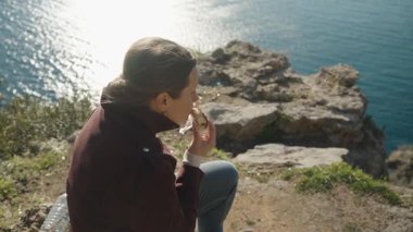 Uçurumun kenarında oturmuş Çikolata kaplı abur cubur yerken Altında Parlayan Okyanus 'a bakan kadın