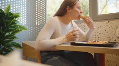 Beyaz kazaklı bir kadın elinde pipetle soğuk bir içecek tutarken pizzayı ısırıyor büyük bir penceresi ve arka planda iç mekan bitkileri olan bir lokanta masasında oturuyor.