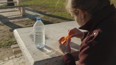 Dışarıdaki parkta bir şişe suyla beton bir masada mandalina soyan kadın.