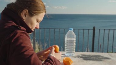 Burgundy ceketli kadın Açık Masa 'da portakal soyuyordu. Güneş ışığıyla Gölgeleri Yüze ve Yüze Yapıyordu. Arka planda Mavi Gökyüzü ile Yakın Çekim