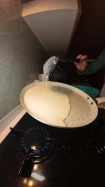 Dikey video. Bir kepçeden bir kepçeye krep hamuru döken kadın Benzin ocağının üzerine bile serpiştirilmiş.