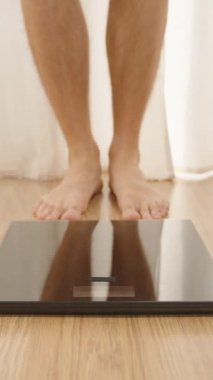 Dikey video. Ahşap zemin üzerinde, Arkaplanda Perdeleri olan Dijital Ölçek Önünde Çıplak Ayaklı Kişi