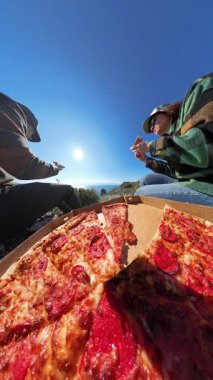 Dikey video. Açık havada pizza yiyen iki arkadaş, manzaralı dağ manzaralı ve okyanus manzaralı, Parlak mavi gökyüzünün altındaki kayalarda oturan, doğada rahatlatıcı bir anın tadını çıkaran iki arkadaş.