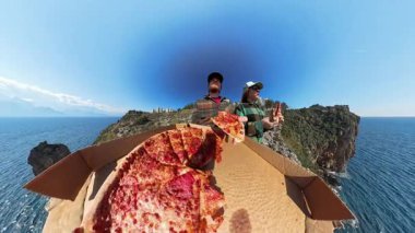 Uçurumun kenarındaki açık bir kutudan Pepperoni Pizza yiyen iki kişi, çarpıcı bir okyanus manzarası olan Parlak Mavi Gökyüzü Altında Geniş Açı Bakış Açısı ile yakalandılar.