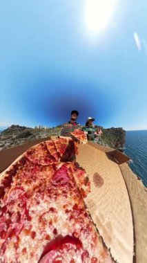 Dikey video. Uçurumun kenarındaki açık bir kutudan Pepperoni Pizza yiyen iki kişi, çarpıcı bir okyanus manzarası olan Parlak Mavi Gökyüzü Altında Geniş Açı Bakış Açısı ile yakalandılar.