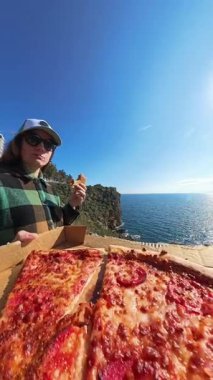 Dikey video. Yeşil Flannel ve Cap giymiş bir kadın okyanusa tepeden bakarken pizza yerken elinde parlak mavi gökyüzünün altında bir kutu biberli pizza tutuyor.
