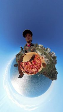 Dikey video. Küçük Gezegen Efektli Bir Pizza Kutusu Tutan Adam, Arka planda Eşsiz 360 derecelik Okyanus, Kayalıklar ve Binalar Görünümü Oluşturuyor