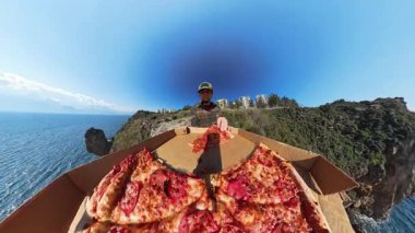 Ön planda Büyük Açık Pizza Kutusuyla Uçurumun Üzerinde Duran Adam, Arkaplanda Geniş Açı Efekt ve Panoramik Okyanus Görünümü ile Yakalandı