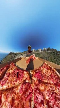 Dikey video. Ön planda Büyük Açık Pizza Kutusuyla Uçurumun Üzerinde Duran Adam, Arkaplanda Geniş Açı Efekt ve Panoramik Okyanus Görünümü ile Yakalandı