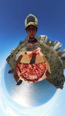 Dikey video. Minik Gezegen Efektli Bir Pizza Kutusu Tutan Adam, Arka planda Eşsiz 360 derecelik Okyanus, Kayalıklar ve Şehir Binaları Görünümü Oluşturdu