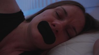 Yatakta uyuyan bir kadın rehine bandıyla nefes alınabilir ağız bandıyla uyku terapisi için düşük ışıktaki siyah şeridi kullanıyor.