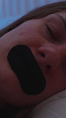 Dikey video. Yatakta uyuyan bir kadın rehine bandıyla nefes alınabilir ağız bandıyla uyku terapisi için düşük ışıktaki siyah şeridi kullanıyor.