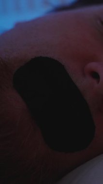 Dikey video. Derin Burun Solunumu Konsepti İçin Loş Mavi Işıkta Yastığa Bantlı Uyuyan Adam 'ın Yakın Çekimi