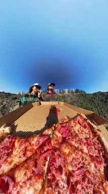 Dikey video. Erkek ve Kadın Uçurumun kenarında Pizzanın Tadını Çıkarırken, Önde Açık Kutu, Geniş Açı Mercek ve Çarpıcı Kıyı Manzarası ile Yakalandı.