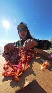 Dikey video. Güneş Gözlüklü Kadın Bir Kutudan Pizza Dilimi Alırken Diğer İki El Uzanıyor, Parlak Güneş Altında Sahne Okyanusu Manzarası