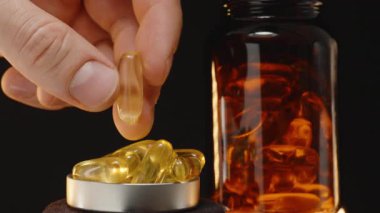El, arka planda şeffaf kahverengi cam bir şişe ile jel kapsüllerle dolu bir metal kapsülden Omega 3 kapsülü alıyor.