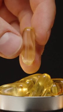 Dikey video. El, arka planda şeffaf kahverengi cam bir şişe ile jel kapsüllerle dolu bir metal kapsülden Omega 3 kapsülü alıyor.