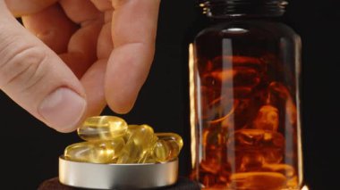 El, Omega 3 kapsülünü jel kapsülleriyle dolu bir metal kapsüle düşürüyor. Arkaplanda şeffaf kahverengi bir şişe var. Karanlık bir ayara karşı.