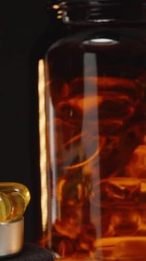 Dikey video. El, Omega 3 kapsülünü jel kapsülleriyle dolu bir metal kapsüle düşürüyor. Arkaplanda şeffaf kahverengi bir şişe var. Karanlık bir ayara karşı.